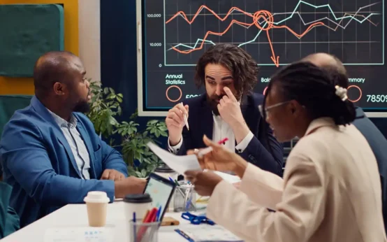 Reunião de empresários analisando gráficos de economia e discutindo estratégias empresariais diante de mudanças econômicas.