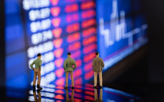 Três figuras de miniatura observando uma tela de mercado financeiro com ações em queda e gráficos, simbolizando estratégias empresariais em cenários de crise econômica.
