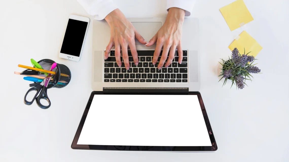 Pessoas trabalhando em um notebook sobre uma mesa branca, com celular ao lado e materiais de escritório, destacando a criação e otimização de landing pages. Landing pages: Como criar páginas que transformam visitantes em leads.