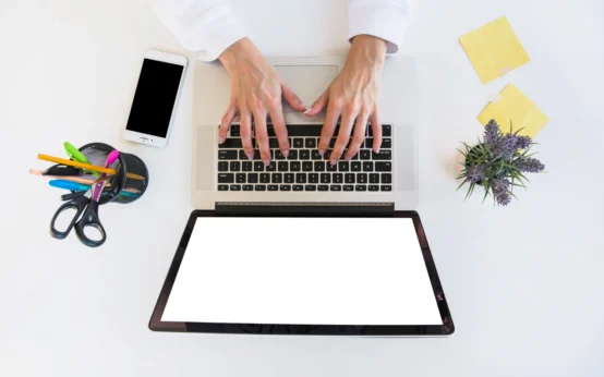 Pessoas trabalhando em um notebook sobre uma mesa branca, com celular ao lado e materiais de escritório, destacando a criação e otimização de landing pages. Landing pages: Como criar páginas que transformam visitantes em leads.