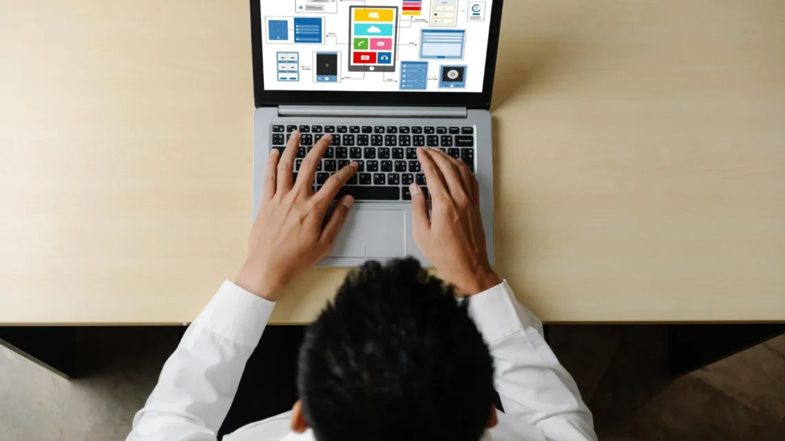 Pessoa trabalhando em notebook com telas de sites e informações empresariais ao fundo, em ambiente de escritório. Site Institucional ou Loja Virtual: Qual a Melhor Opção para Cada Empresa?