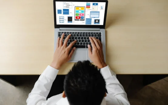 Pessoa trabalhando em notebook com telas de sites e informações empresariais ao fundo, em ambiente de escritório. Site Institucional ou Loja Virtual: Qual a Melhor Opção para Cada Empresa?