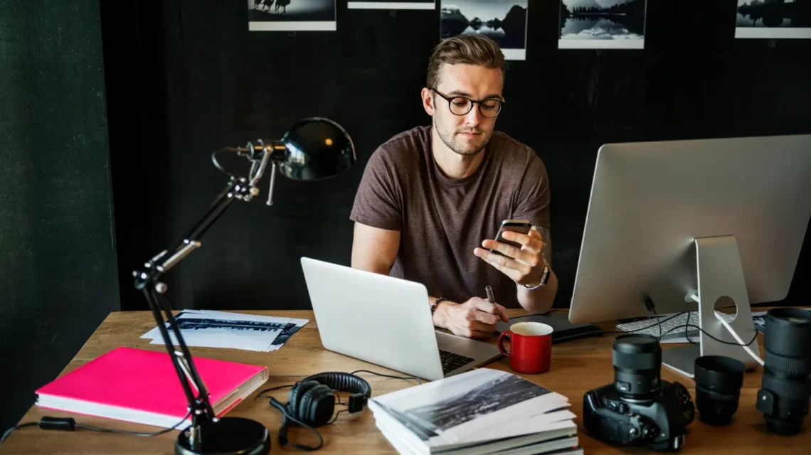 Homem trabalhando em estúdio com notebook e celular, segurando o telefone enquanto usa computador; ao redor há câmera, lentes, microfone e materiais de criação, sugerindo conteúdo para a creator economy em transição: por que executivos de mídia estão migrando de agências para plataformas de criadores.