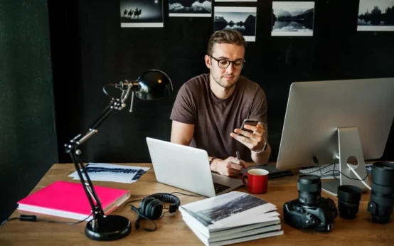Homem trabalhando em estúdio com notebook e celular, segurando o telefone enquanto usa computador; ao redor há câmera, lentes, microfone e materiais de criação, sugerindo conteúdo para a creator economy em transição: por que executivos de mídia estão migrando de agências para plataformas de criadores.